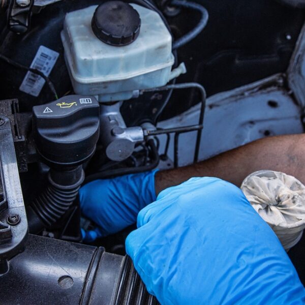 Auto mechanic wearing blue gloves holding a dirty car coolant reservoir filter, inspecting engine parts for maintenance and repair near an open vehicle hood. Fixing vehicle cooling system.