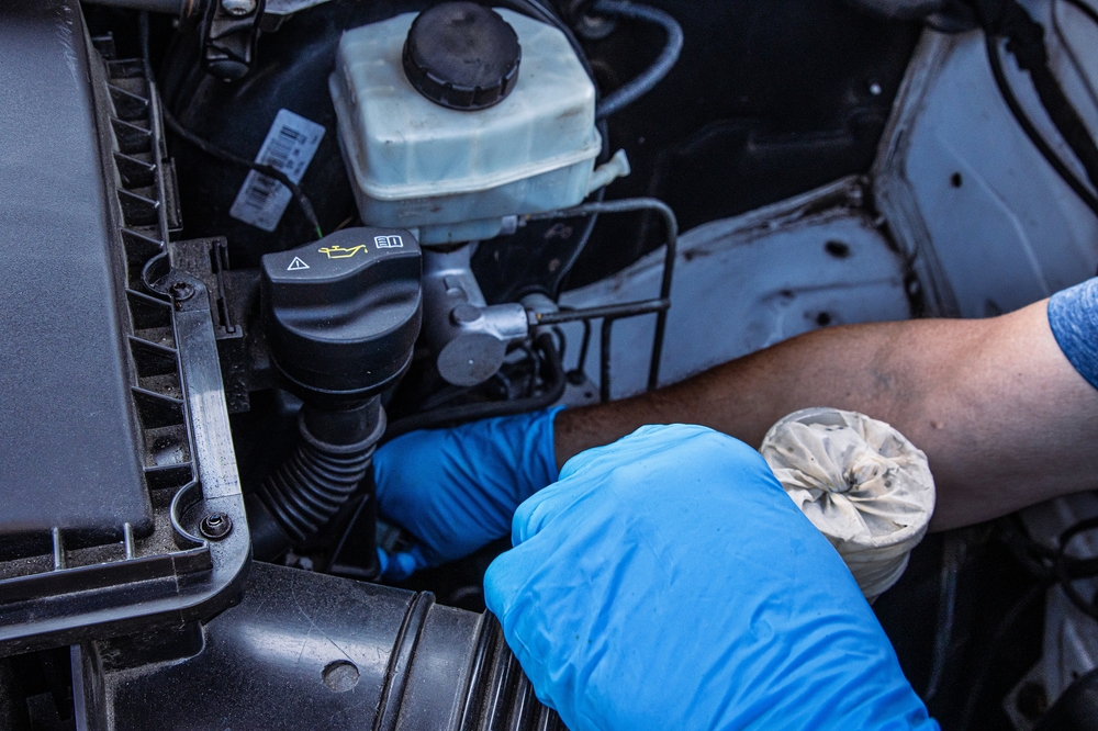 Auto mechanic wearing blue gloves holding a dirty car coolant reservoir filter, inspecting engine parts for maintenance and repair near an open vehicle hood. Fixing vehicle cooling system.